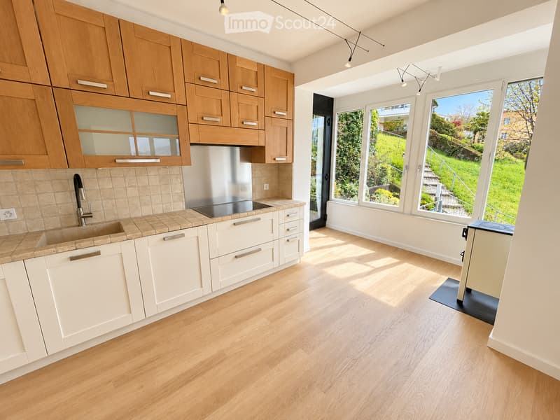 Modern kitchen with light wooden cabinets, white lower cabinets, a central island, and large windows overlooking a green garden.