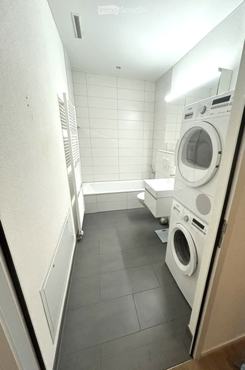 Modern bathroom with white tiled walls, dark grey tiled floor, bathtub, toilet, sink, and stacked washing machine and dryer unit.