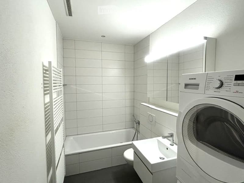 Modern bathroom with white tiled walls, bathtub, sink, toilet, mirror, and a washing machine.