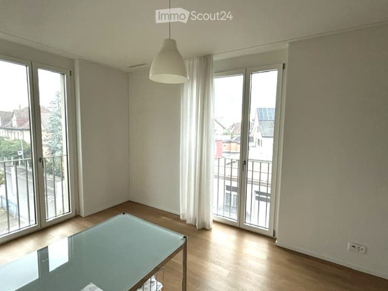 Bright, unfurnished room with large windows and a balcony. Features a modern glass table and wooden flooring.