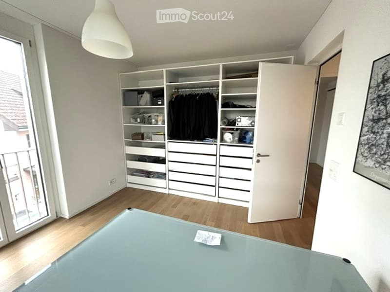 Modern, minimalist room with a large, open wardrobe, a sleek glass table, and a balcony view. The room features light wooden flooring and white walls.