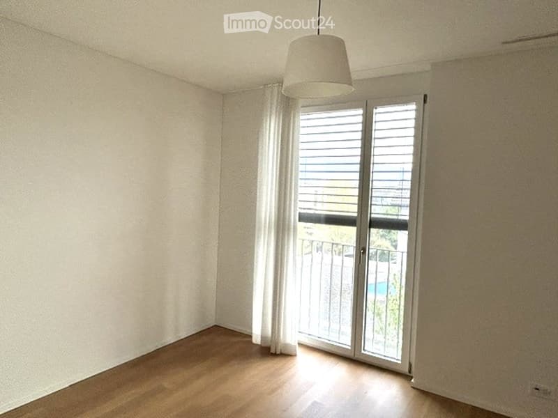 Empty room with wooden flooring, white walls, large window with blinds, and a hanging lamp.