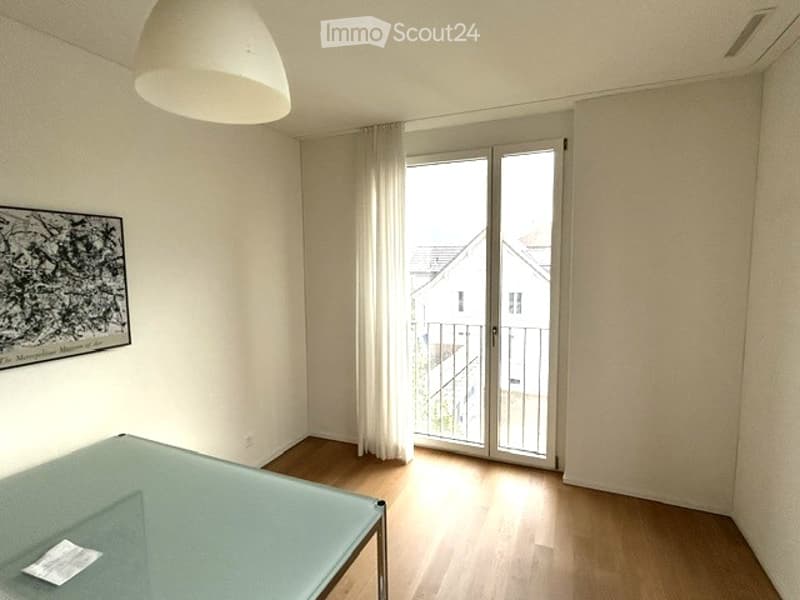 Empty room with wooden flooring, large sliding glass door leading to a balcony, white walls, and a modern pendant light fixture.