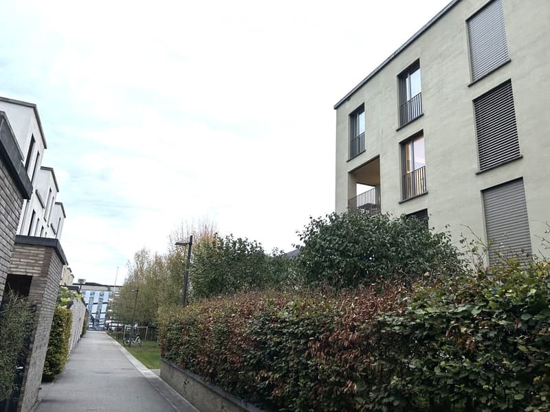 Modern multi-story residential building with balconies and large windows, surrounded by greenery and a paved walkway.