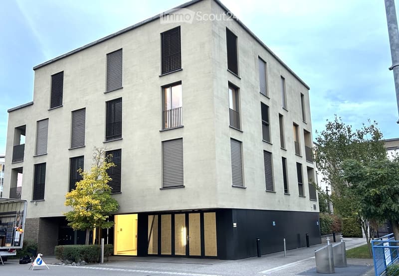 Modern multi-story building with a light beige facade, large windows, and balconies. The ground floor features a black entrance area with glass doors and a small tree in front.