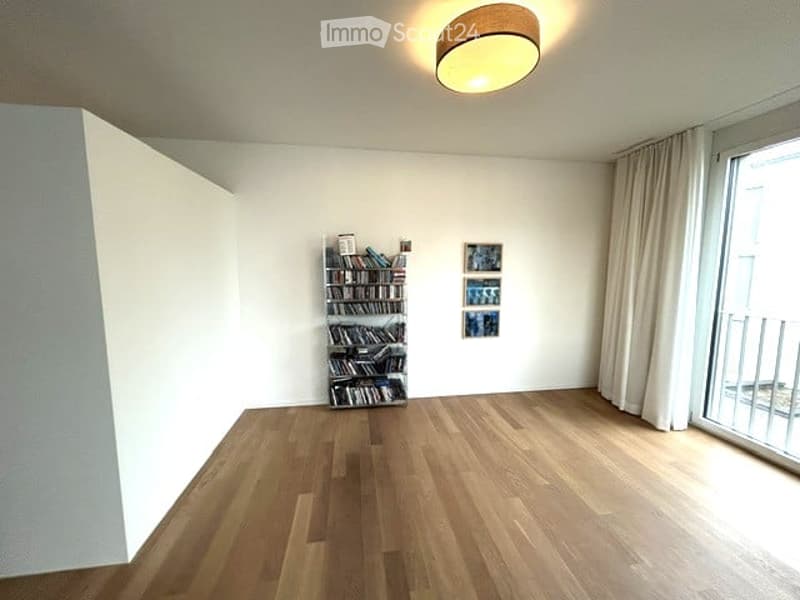 Bright, unfurnished room with wooden flooring, large window with white curtains, and a small bookshelf with books and picture frames on the wall.