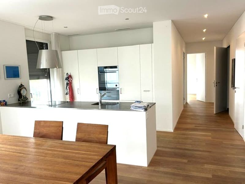 Modern, open-plan kitchen and dining area with white cabinets, a wooden dining table, and chairs. The kitchen features a sink, faucet, and oven, with a lamp hanging above the counter.