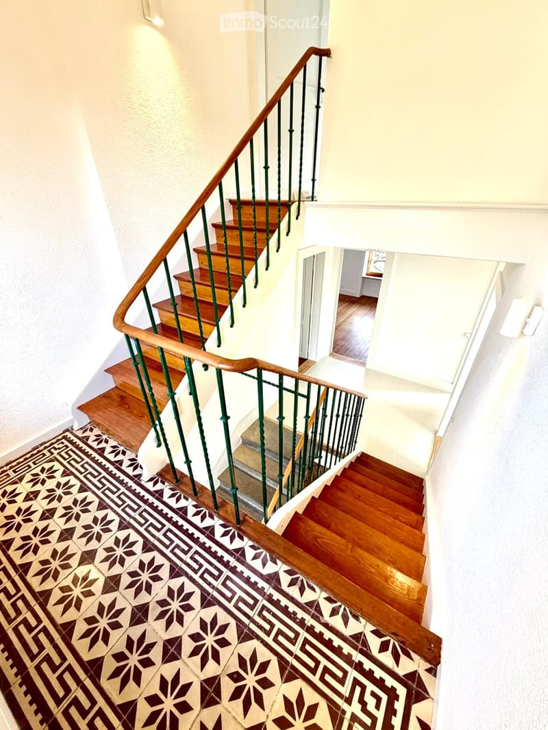 Spiral staircase with wooden steps and green metal railing, patterned tile floor, white walls, and warm lighting.