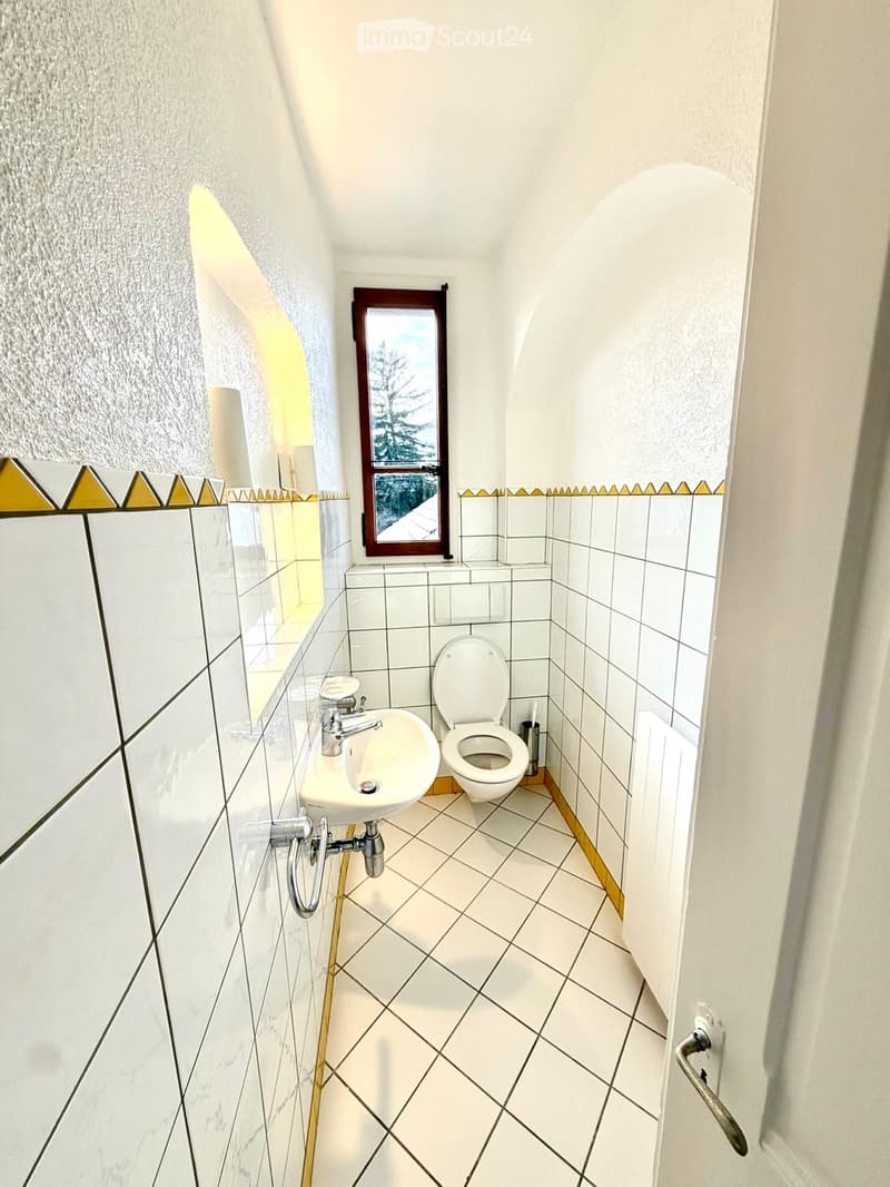 Small bathroom with white tiled walls and floor, a sink with a chrome faucet, a toilet, and a mirror with a decorative border.