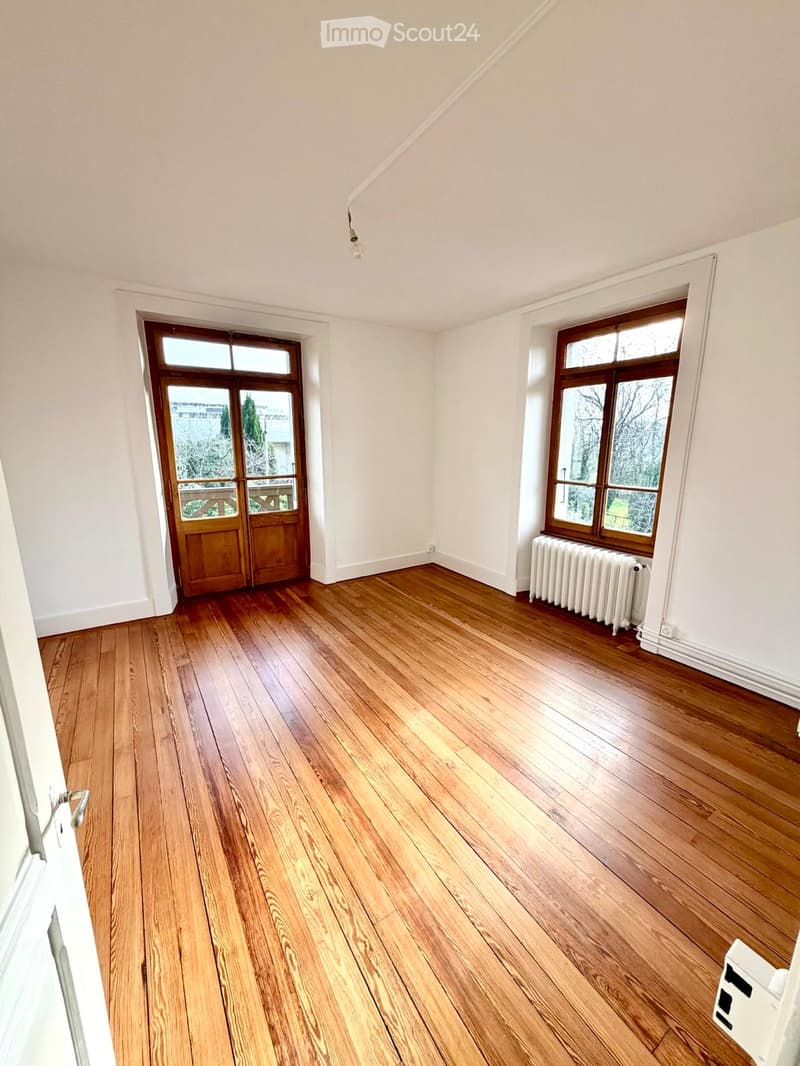 Empty room with wooden flooring, white walls, large windows, and a radiator. The room has a wooden door leading outside.