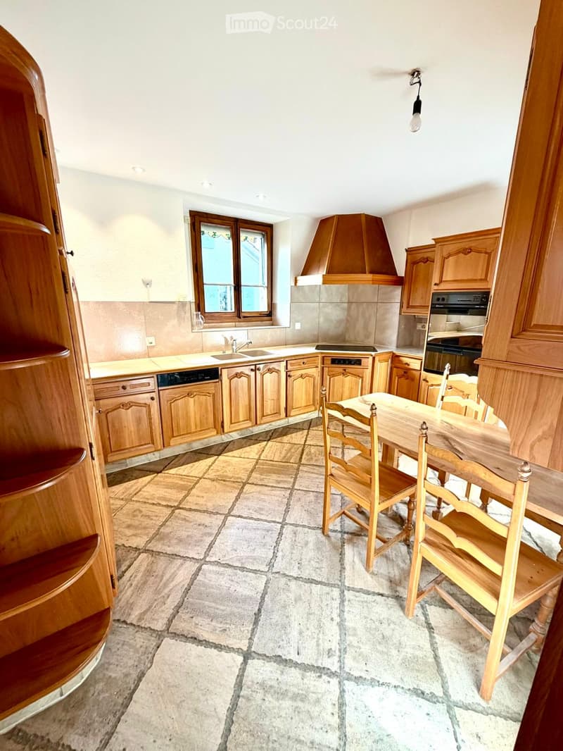 A well-lit kitchen with wooden cabinets, a central island, a dining area with a wooden table and chairs, and a tiled floor.