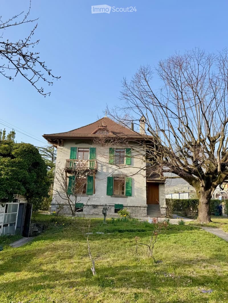 Two-story house with a tiled roof, green shutters, and a balcony on the upper floor. The house is surrounded by a well-maintained lawn and trees.