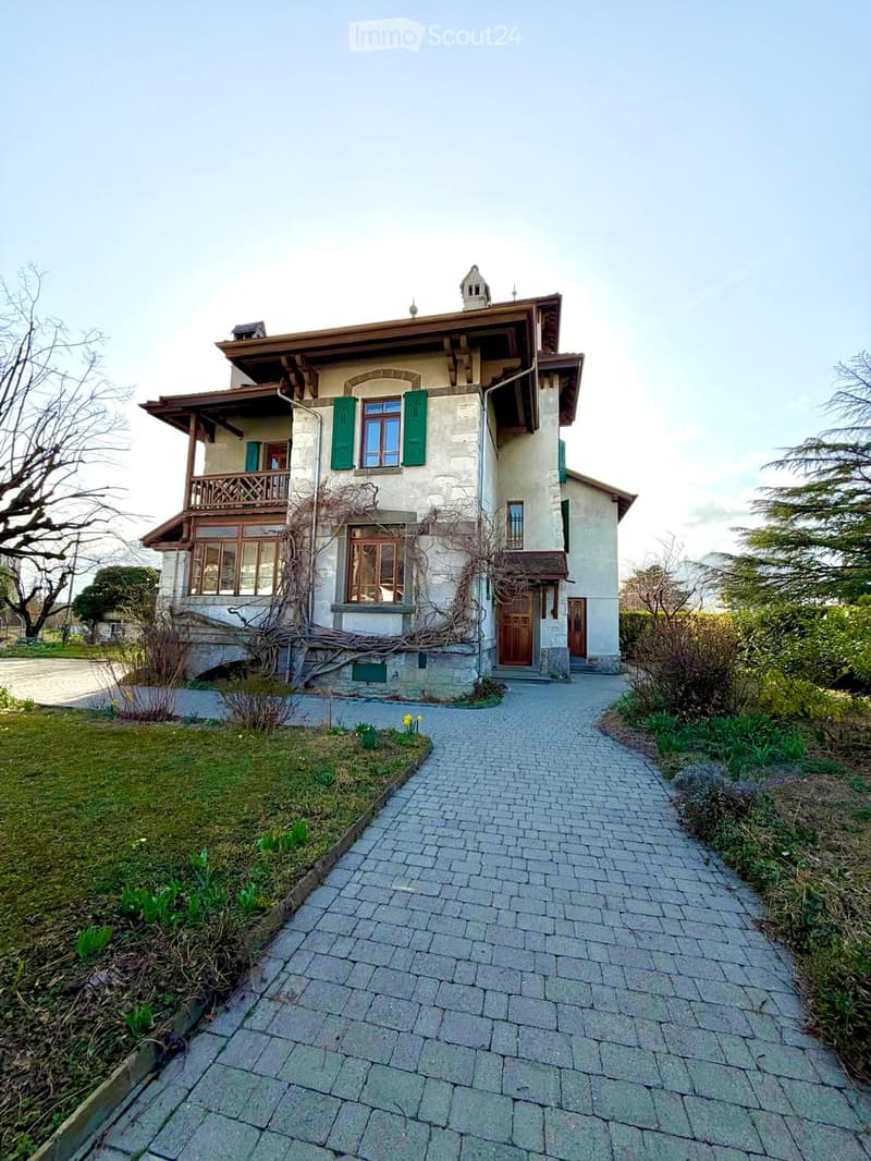 Two-story house with beige stone facade, green shutters, wooden balcony, and a brick pathway leading to the entrance.