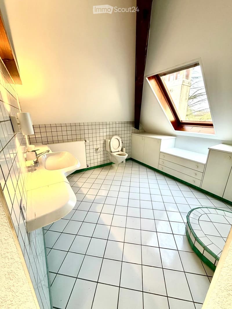 Bathroom with white tiled floor and walls, a sink, toilet, and built-in cabinet. Features a skylight window and a small shelf.