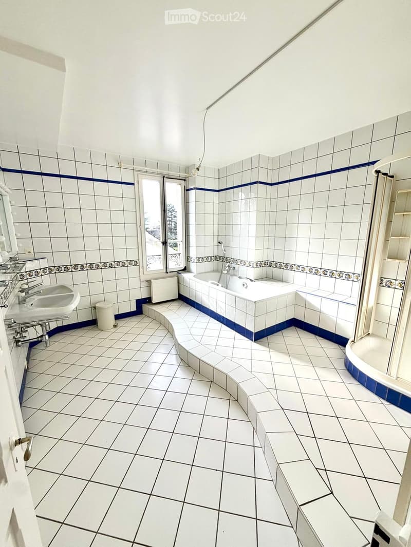 Bathroom with white tiled walls and floor, blue decorative borders, a bathtub, a sink, a radiator, and a window with a view.