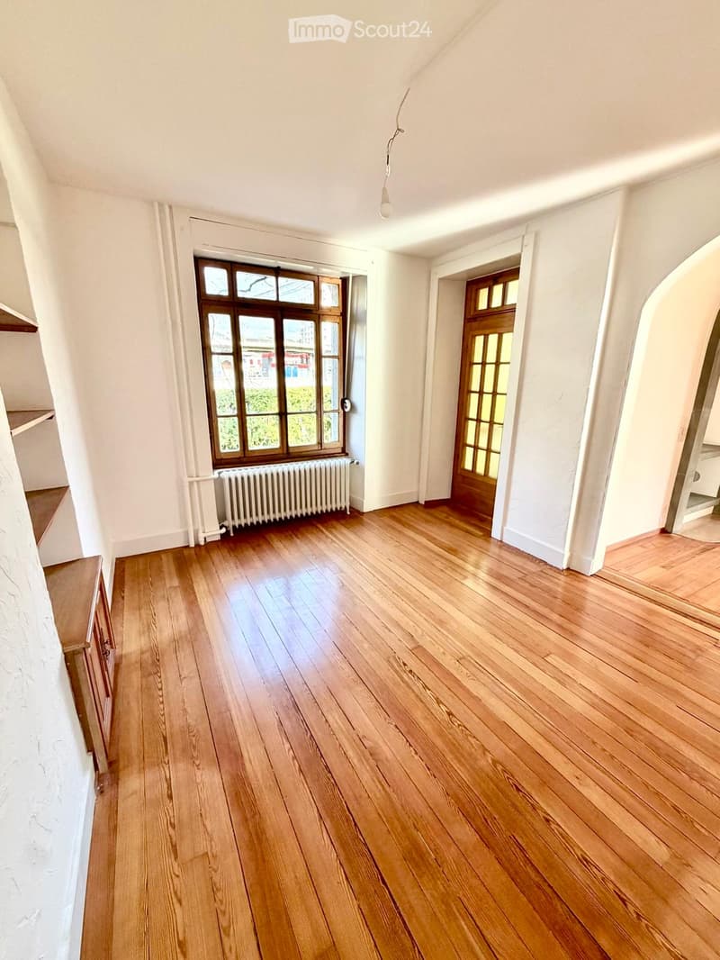 Empty room with wooden flooring, white walls, large window with wooden frame, radiator, and a wooden door with glass panels.