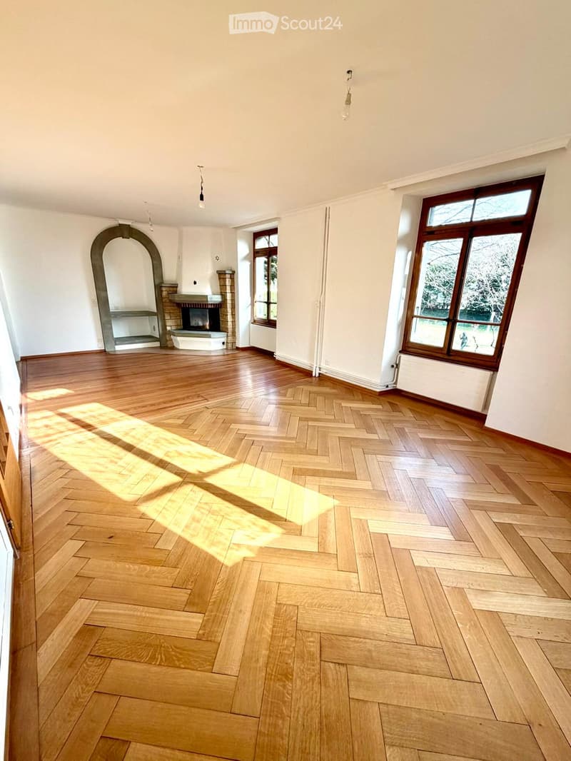 Empty room with wooden herringbone flooring, white walls, large windows, and a fireplace with a stone surround.