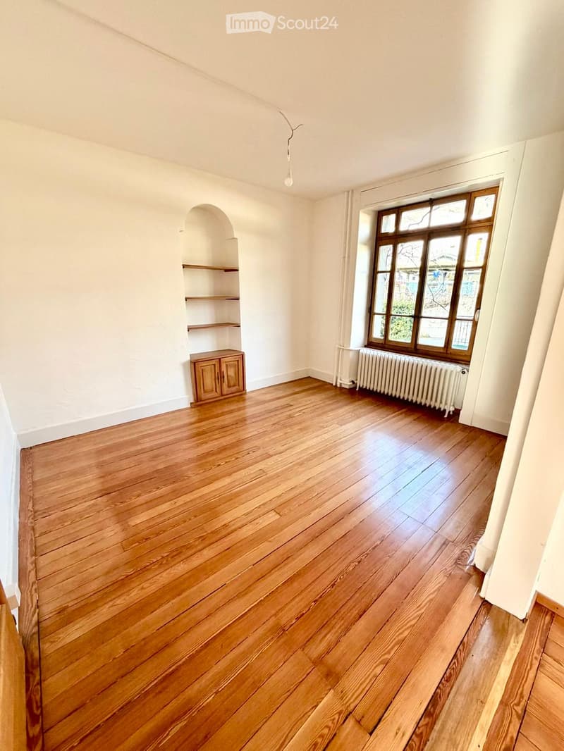 Empty room with wooden flooring, white walls, a window with wooden frames, a radiator, and a small wooden cabinet with shelves.