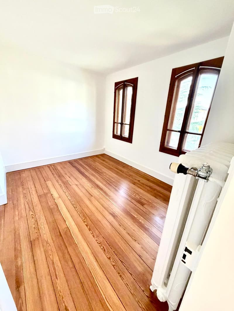 Empty room with wooden flooring, white walls, and two windows with wooden frames. A white radiator is visible on the right side.