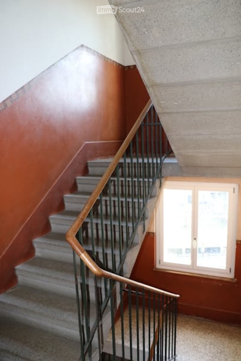 The image shows a staircase leading up to an upper floor. The staircase has a wooden handrail and metal steps. The wall on the left side of the staircase is painted in a brown color, while the wall on the right side is white.