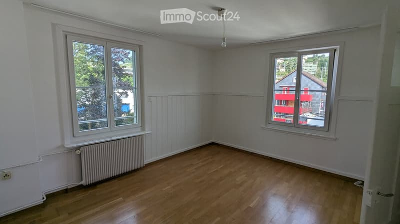 Empty room with wooden flooring, white walls, and two large windows offering a view of the outside. There is a radiator under one window and a light fixture hanging from the ceiling.