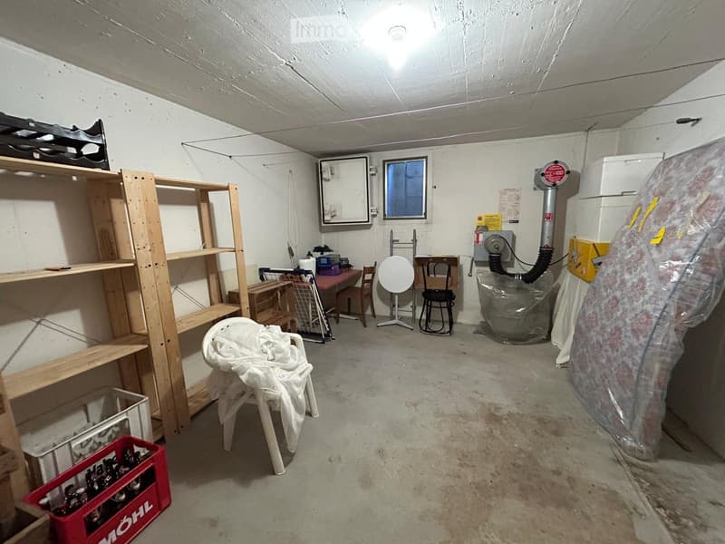 The room is a basement or utility space with concrete floors and white walls. It contains a wooden shelving unit, a small table with chairs, a plastic chair with a cloth draped over it, and various storage items including a red crate and a large covered o