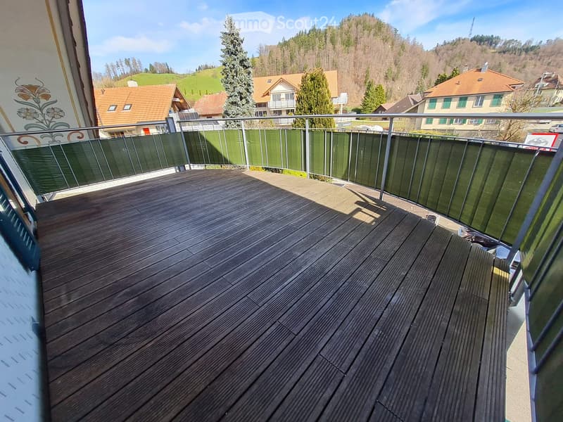 Wooden deck with metal railing, green privacy screen, overlooking a hilly landscape with houses and trees.