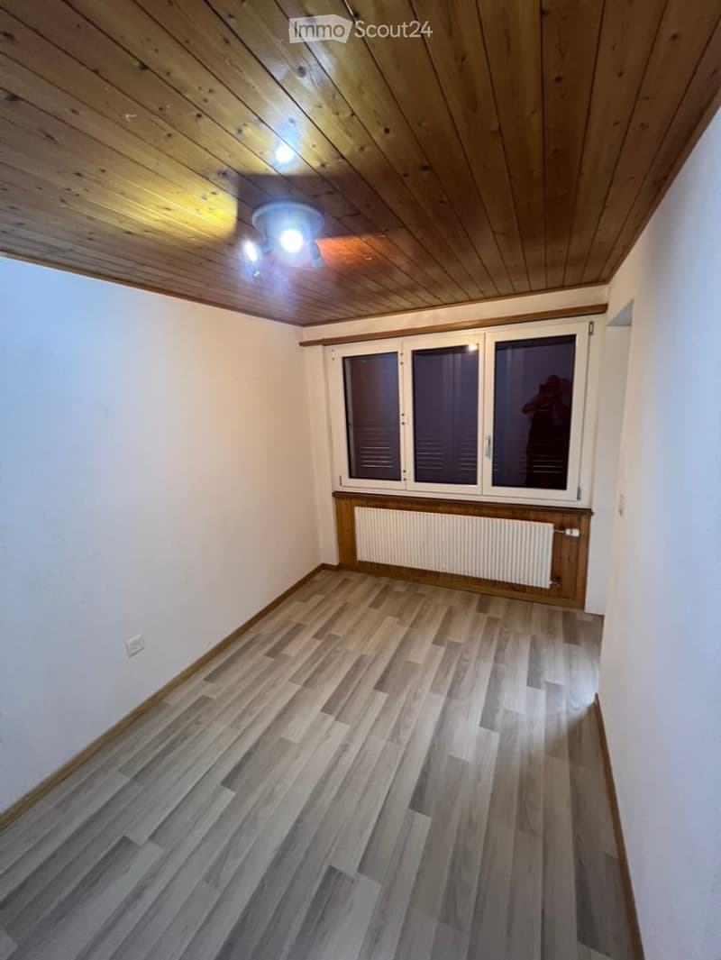 Empty room with wooden flooring, white walls, and a large window with blinds. The ceiling is wooden with recessed lighting.