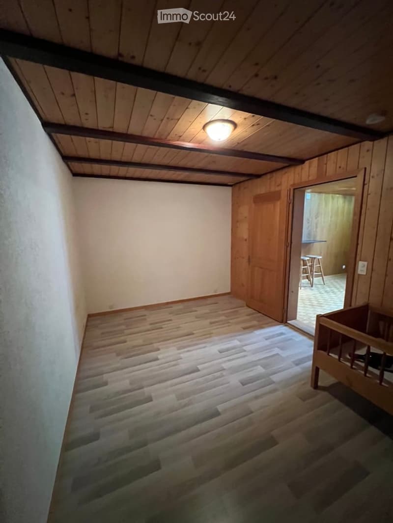 Empty room with wooden walls, exposed wooden beams on the ceiling, and a wooden floor. An open doorway leads to another room with visible furniture.