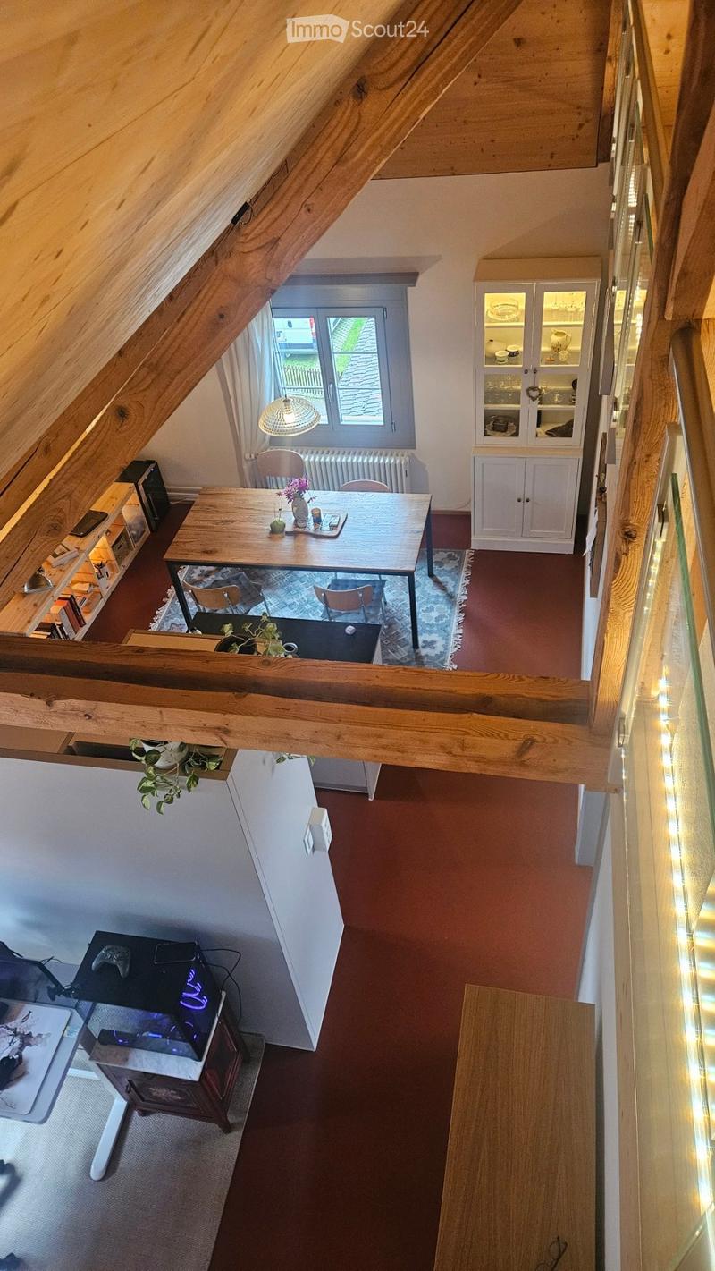 Loft-style room with wooden beams, a dining table with chairs, a cabinet, a lamp, and a gaming setup with a screen and plant.