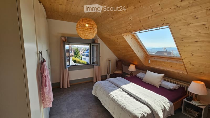 Cozy attic bedroom with wooden walls and ceiling, large skylight and window offering a scenic view, featuring a double bed, nightstands, lamps, and a chair.