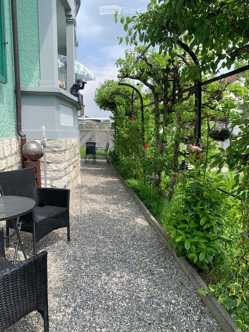 Garden path lined with gravel, featuring lush greenery, a small seating area with a table and chairs, and a decorative statue on the building.