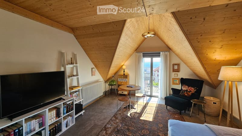 Cozy attic living room with wooden beams, large window with view, sofa, armchair, coffee table, floor lamp, bookshelf, and television.