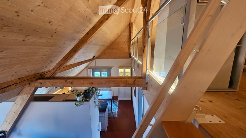 A modern, open-concept hallway with exposed wooden beams and a slanted ceiling. The space features a staircase leading to an upper level, with a view of a dining area below that includes a table, chairs, and potted plants. The walls are painted in a neutr