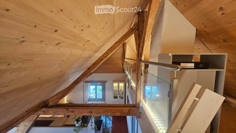 A modern, wooden staircase with glass railings leads to an open-plan living area below. The ceiling features exposed wooden beams, and the walls are painted in a neutral tone. The lower level shows glimpses of furniture and decor, including a potted plant