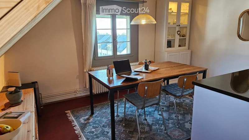 A cozy, well-lit room with a wooden table, laptop, chairs, a hanging lamp, and a window view. The room features a patterned rug, a small cabinet, and a bowl of fruit on a side table.
