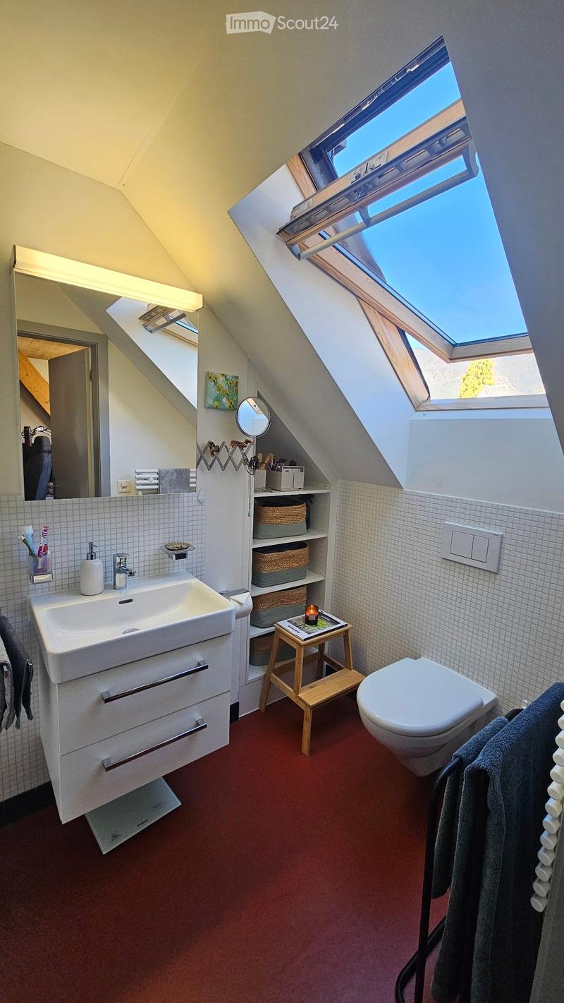Modern attic bathroom with skylight, white tiled walls, red carpet, sink, toilet, small wooden stool, mirror, and shelves with baskets.