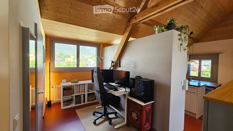 Cozy attic workspace with wooden beams, large windows offering a scenic view, a desk with dual monitors, a comfortable chair, and a small sink area.