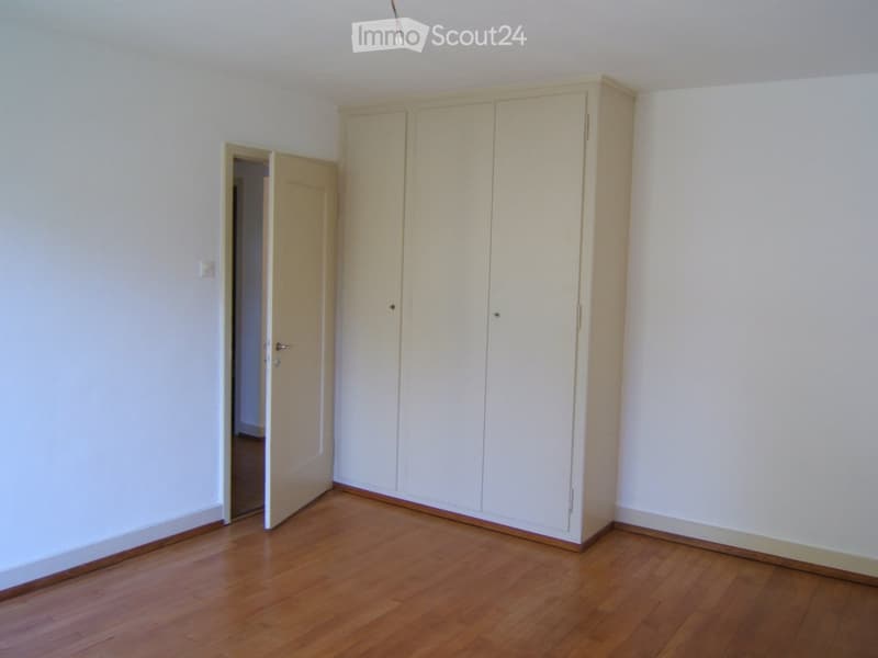 Empty room with light-colored wooden flooring, white walls, and a large built-in wardrobe unit. A door is visible on the left side leading to another space.