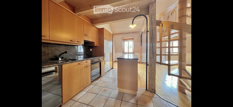 Bright kitchen with wooden cabinets, stainless steel appliances, and a central island with a stool. The space features a mix of tiled and wooden flooring, and a tall floor lamp provides additional lighting.