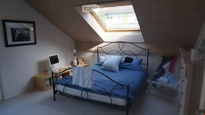 Cozy attic bedroom with slanted wooden ceilings, skylight window, double bed with blue bedding, nightstand with laptop, picture frame on wall, and wardrobe with clothes.