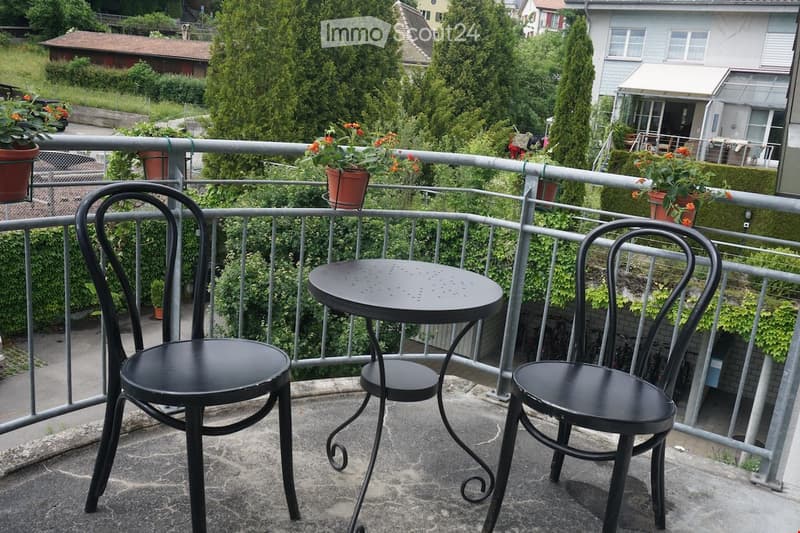 Balcony with a small black table and two black chairs, surrounded by potted plants and greenery.