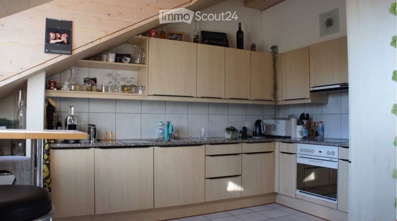 A cozy kitchen with light wooden cabinets, a white oven, and various appliances on the counter, including a coffee maker and a kettle. Shelves above the cabinets hold decorative items and glassware.