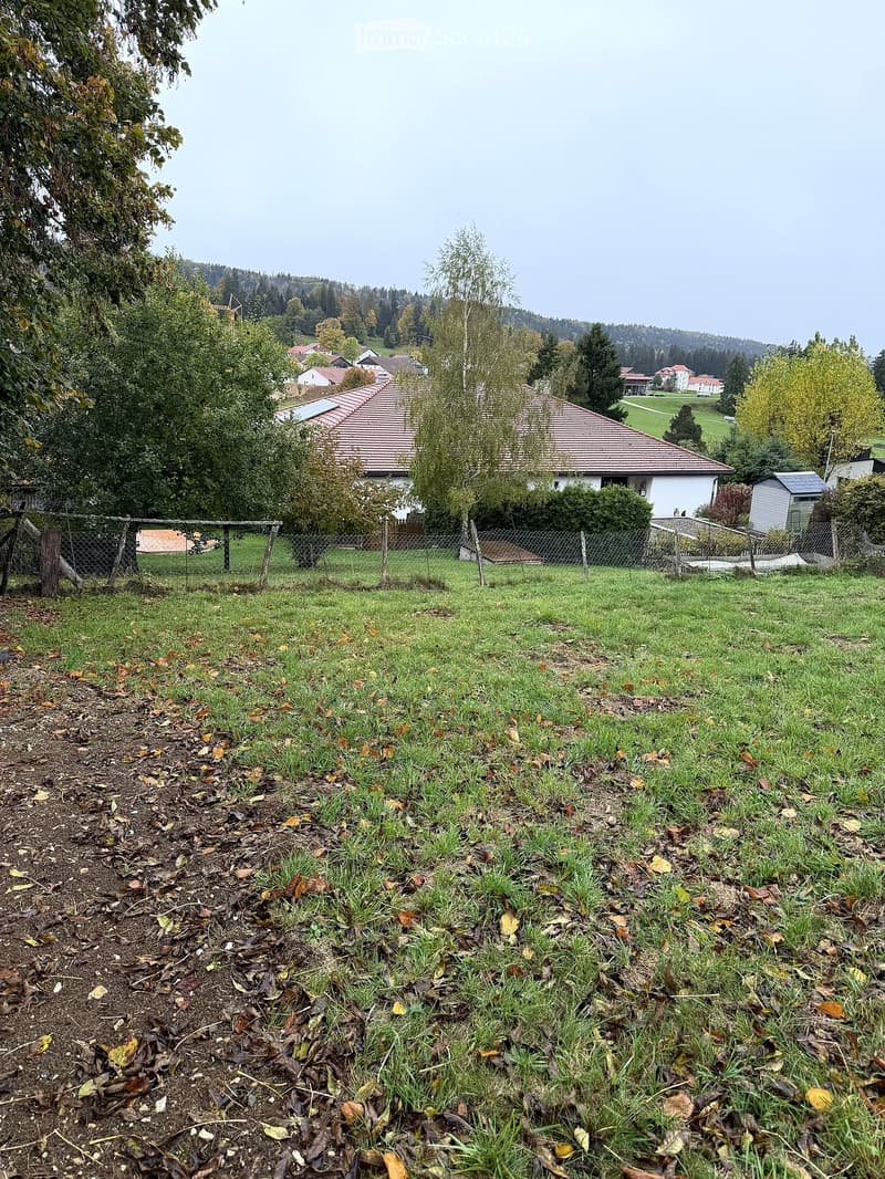 A grassy yard with a fence, trees, and a house in the background, surrounded by a rural landscape with other houses and hills in the distance.