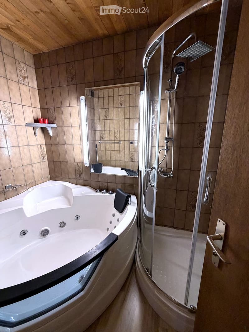 Bathroom with a large bathtub, walk-in shower, and tiled walls. Features include a handheld showerhead, grab bars, and a small shelf with a red cup.