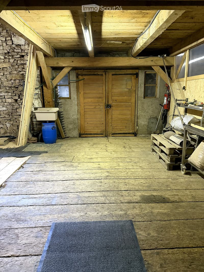 Empty wooden-floored room with exposed wooden beams, stone walls, a sink, a blue barrel, a fire extinguisher, and a wooden door.