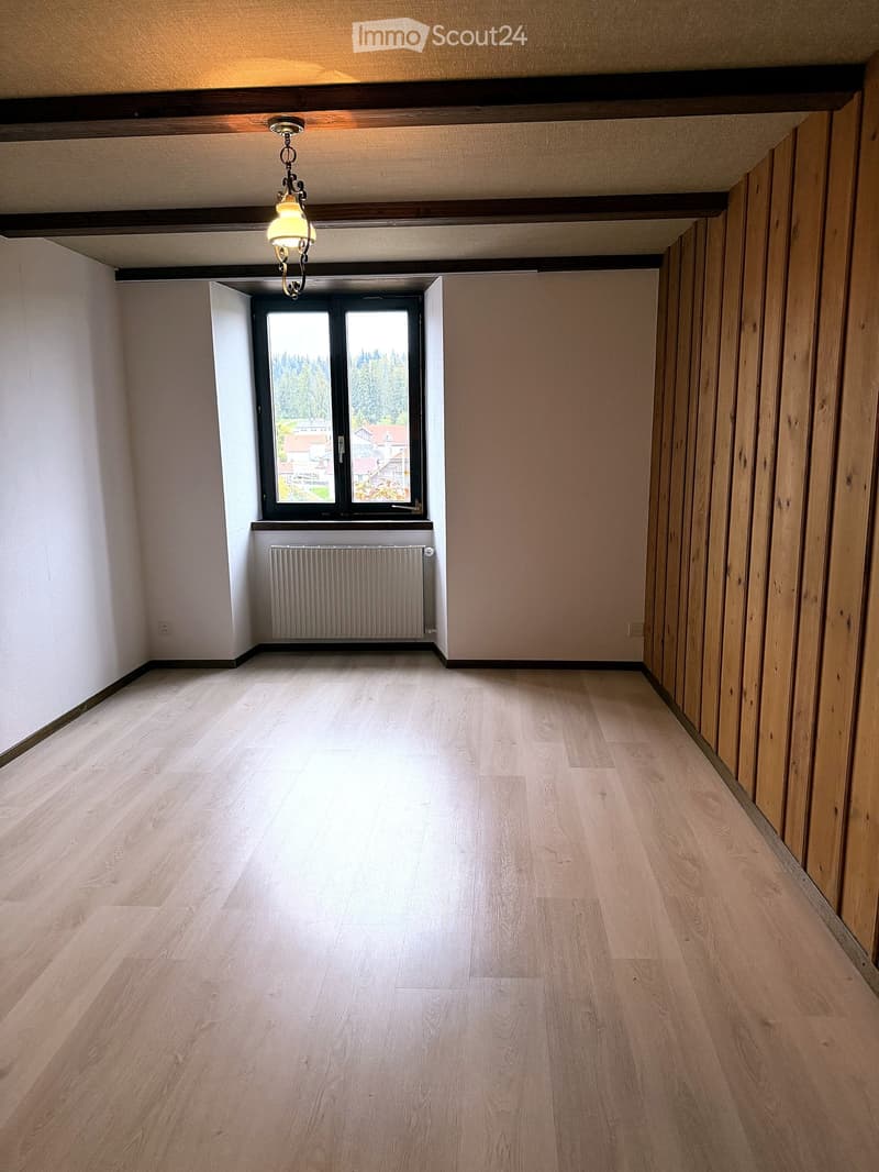 Empty hallway with wooden flooring, white walls, and a window with a radiator below it. The ceiling has exposed wooden beams and a hanging light fixture.