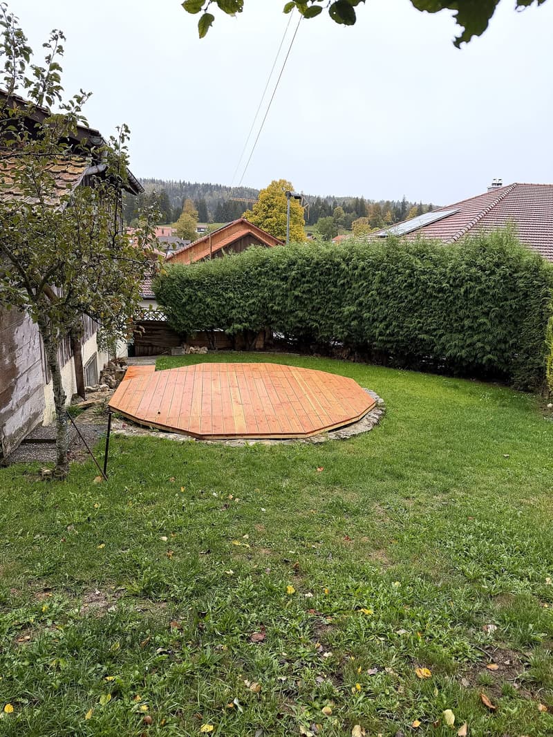 A well-maintained garden with a wooden deck, green lawn, and surrounding trees and shrubs.