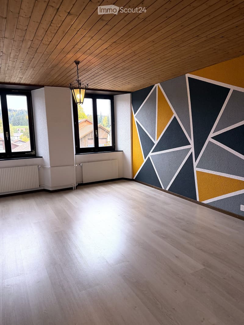Empty room with wooden flooring, white walls, and large windows offering a view of the outdoors. The walls feature a modern geometric design with dark and yellow panels.