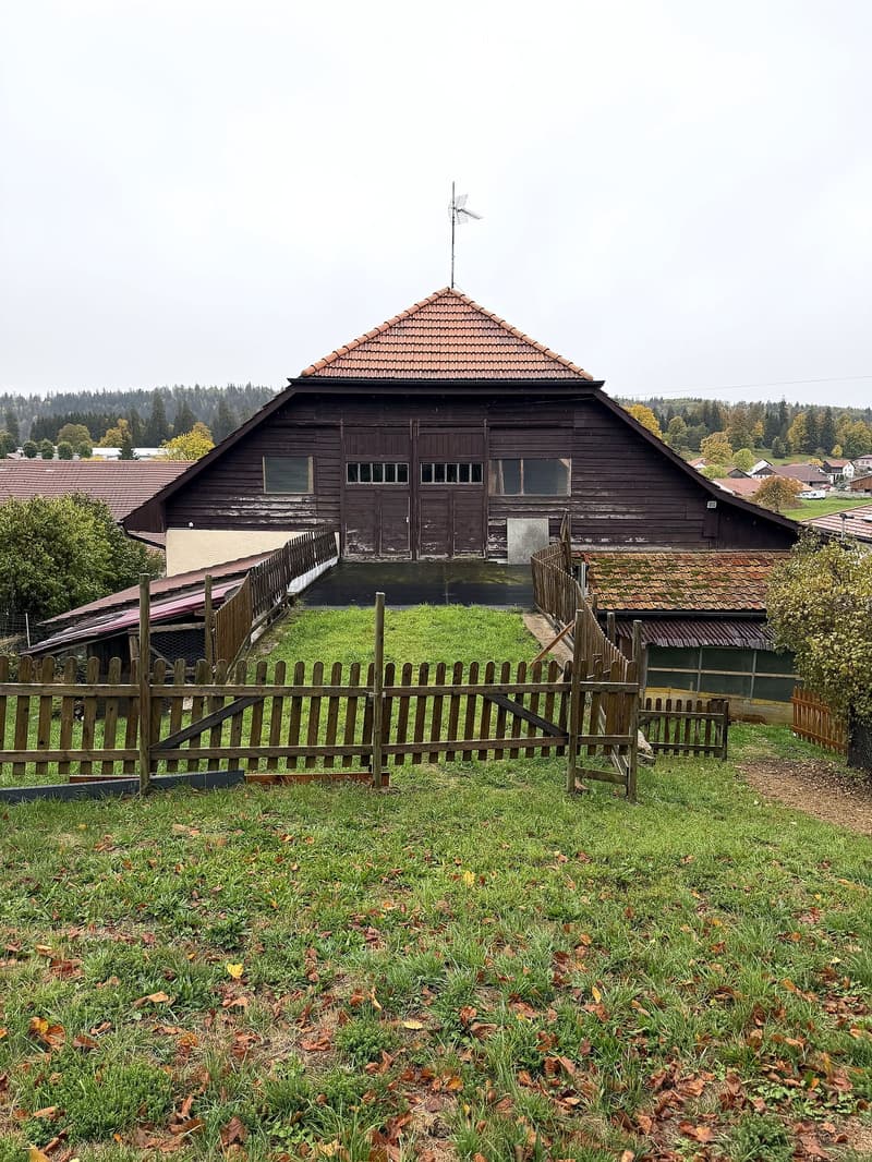 A large wooden barn with a pitched roof and multiple windows, surrounded by a wooden fence and grassy yard.